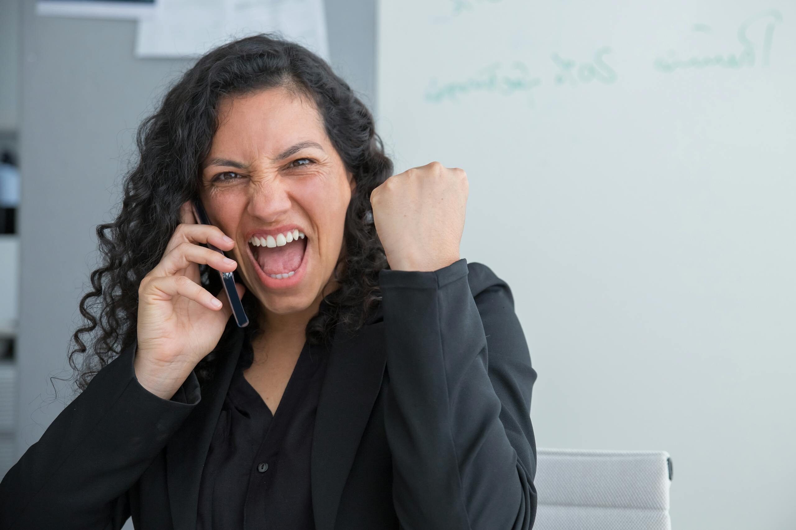 A joyful businesswoman with curly hair celebrates success while talking on the phone indoors.